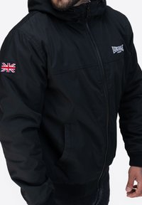 Czarna kurtka z kapturem i zamkiem, z naszywką w stylu Union Jack na rękawie oraz bocznymi kieszeniami. Gładki materiał i ściągacze przy rękawach.