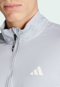 Lichtgrijze sportjack met hoge kraag, rits aan de voorkant en zwarte trekkoord. Bevat een witte Adidas-logo op de borst.