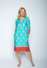 Lekki turkusowy tunika o geometrycznym wzorze w odcieniu kremowym, z czerwonym wzorzonym dołem, dekoltem w serek oraz bocznymi kieszeniami.