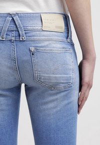 Pantalones vaqueros de mezclilla azul claro con un diseño ajustado. Presenta dos bolsillos traseros, una etiqueta de marca visible y herrajes de metal en la cintura.