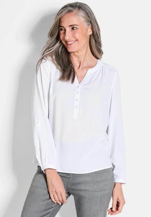Blouse blanche à manches longues avec décolleté en V, patte de boutonnage et épaule froncée. Portée avec un jean gris. Texture de tissu lisse.