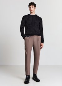 Schwarzer Pullover mit Zopfmuster und Rundhalsausschnitt, kombiniert mit hellbraunen, plissierten Hosen und schwarzen Stiefeletten. Glatte Texturen und maßgeschneiderte Passform.