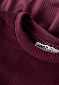 Bordeaux ribbattu puuvillapaita, jossa on pyöreä pääntie ja etiketti, joka osoittaa koon EU 92, US 2T. Tekstuuriltaan pehmeä, näkyvillä urilla.