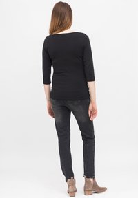 Svart långärmad topp med figursydd design, kombinerad med mörka jeans. Modellen bär taupe ankelstövlar med en staplad klack. En enkel vit bakgrund.