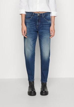 Femme portant un jean bleu délavé taille mi-haute, associé à des bottines noires et une chemise blanche courte à boutons, debout devant un fond uni.