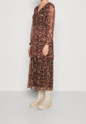 Femme portant une robe midi à manches longues avec un motif floral, une jupe à volants et des bottines épaisses beiges, debout contre un mur uni.