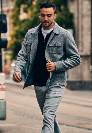 Grijze wolmix jas met twee borstzakken, gedragen over een zwarte zip-up top. Lichtgrijze broek met een textuurafwerking. Stedelijke omgeving.