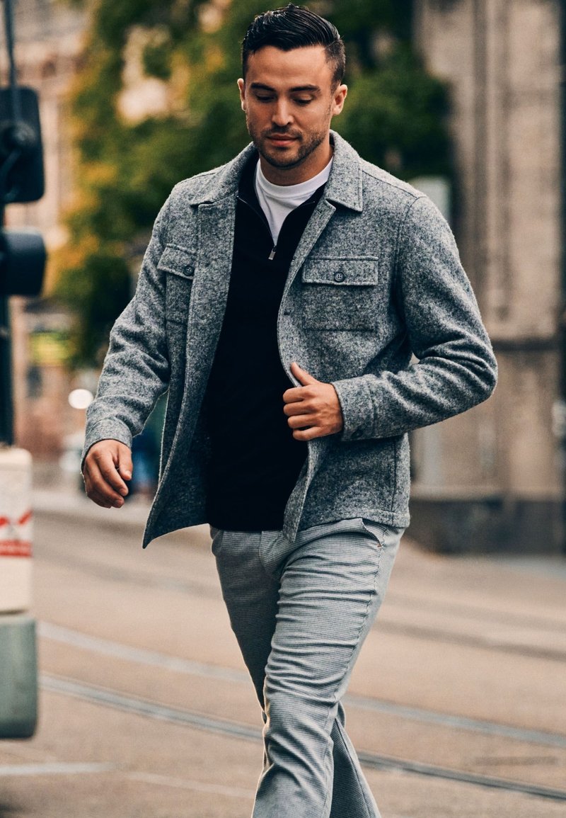 Grijze wolmix jas met twee borstzakken, gedragen over een zwarte zip-up top. Lichtgrijze broek met een textuurafwerking. Stedelijke omgeving.