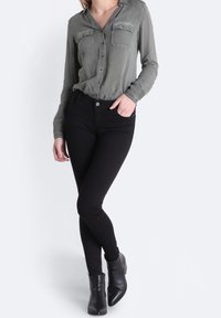 Olivgrön knapp-up skjorta med två bröstfickor, kombinerad med svarta skinny jeans och svarta ankle boots, stående mot en vit bakgrund.