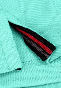 Helles türkisfarbiges Gewebe mit einer strukturierten Oberfläche, das eine diagonale Öffnung aufweist, die eine gestreifte Bordüre in rot, schwarz und grauen Tönen zeigt.