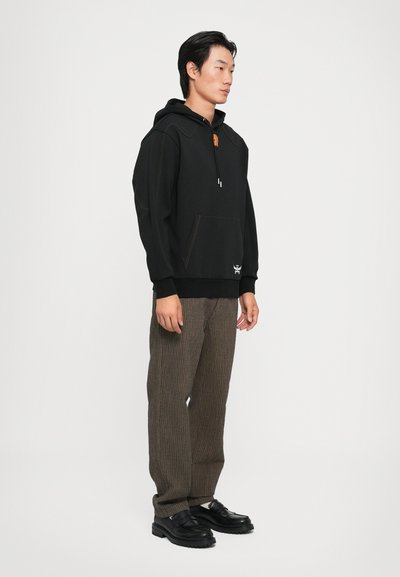 Felpa nera con cappuccio e dettagli arancioni, dotata di tasca frontale, abbinata a pantaloni a righe marroni e scarpe chunky nere.
