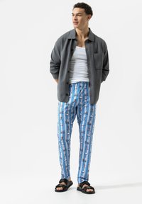 Graues Hemd über einem weißen Tanktop, kombiniert mit blauen Hosen mit Fahrrad- und Roller-Print und schwarzen Pantoletten.