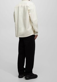 Camisa de botones color crema con un corte relajado, mangas largas y dobladillo curvado, combinada con pantalones negros y zapatillas negras.