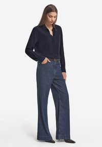 Granatowa koszula z długim rękawem i kołnierzykiem, zestawiona z szerokimi niebieskimi dżinsami. Spodnie mają wykończony brzeg i kieszenie z przodu.