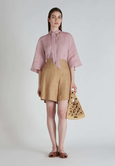 Blusa di lino rosa chiaro con scollo a fiocco, pantaloni corti di lino color mattone e borsa di paglia all'uncinetto. Scarpe marroni senza lacci completano il look.