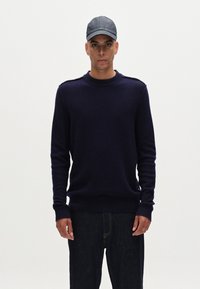 Mörkblå stickad tröja med rund hals och ribbade ärmslut; kombinerad med mörka jeans och en mörk denimkeps. Enkel, strukturerad design.