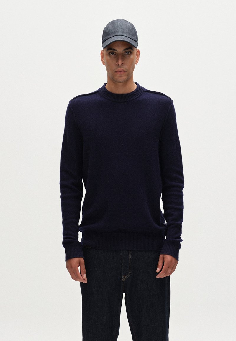 Mörkblå stickad tröja med rund hals och ribbade ärmslut; kombinerad med mörka jeans och en mörk denimkeps. Enkel, strukturerad design.