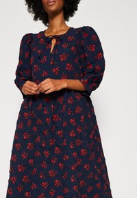 Femme portant une robe bleu marine à imprimé floral rouge, manches bouffantes trois-quarts et encolure goutte d'eau avec lien. Mains jointes à la taille.