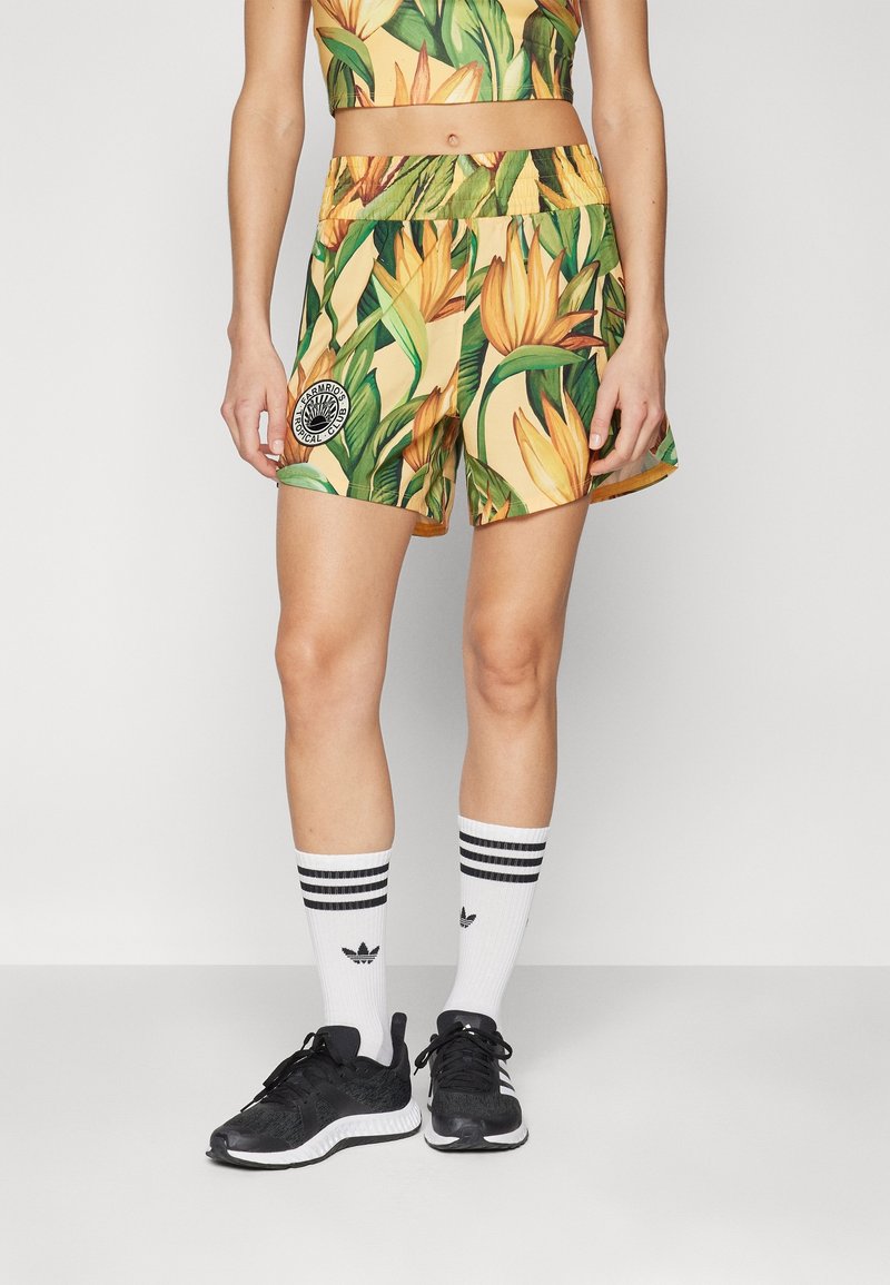 Personne portant un short jaune à imprimé tropical avec un logo circulaire, un crop top assorti, des chaussettes blanches Adidas et des chaussures de course noires.