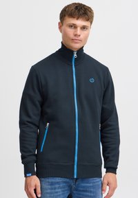 Sort lynlåstrøje med høj krave, blå lynlås og detaljer, to sidelommer og ribstrikkede manchetter, lavet af tekstureret stof.