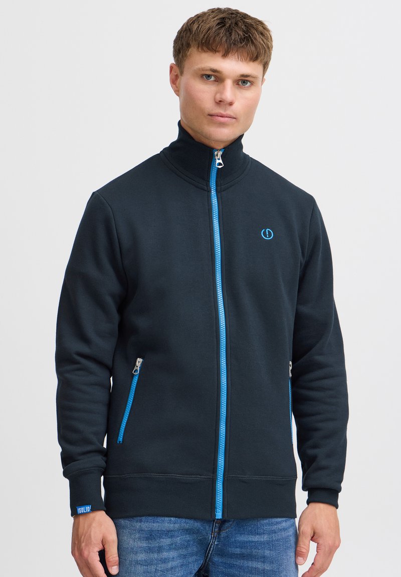 Sort lynlåstrøje med høj krave, blå lynlås og detaljer, to sidelommer og ribstrikkede manchetter, lavet af tekstureret stof.