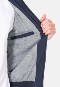 Giacca a zip blu navy con interior grigio, dotata di una piccola tasca interna e polsini a costine per una vestibilità aderente. Testura esterna liscia.