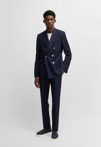Navy double-breasted blazer with silver buttons, paired with matching slim-fit trousers and navy slip-on shoes, featured against a neutral background.