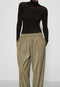 Svart långärmad polotröja i genomskinligt material, kombinerad med ljus olivgrön, plisserade, högmidjade byxor med elastisk midja.