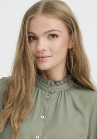 Green blouse with ruffled collar, lightweight material, and button details. Hair styled with soft waves, showcasing a natural look.