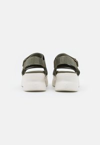 Zelené sandály s nastavitelnými popruhy, silnými bílými podrážkami a popisky na popruhu; otevřený zadní design pro snadné obouvání.