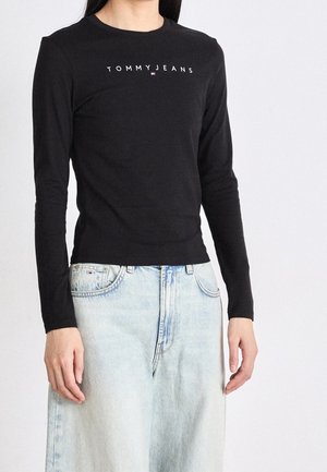 Osoba ubrana w czarną koszulkę z długim rękawem z logo "TOMMY JEANS" i małą flagą, w połączeniu z jasnoniebieskimi jeansami z wysokim stanem.