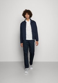 Jaqueta leve azul marinho com dois bolsos frontais, usada sobre uma camisa polo branca. Combinada com calças escuras e tênis brancos.