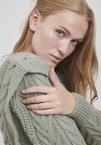 Dicker Strickpullover in sanftem Grün, mit Kabelmustern und gerippten Bündchen. Modell mit langen Haaren, das Textur und Details präsentiert.