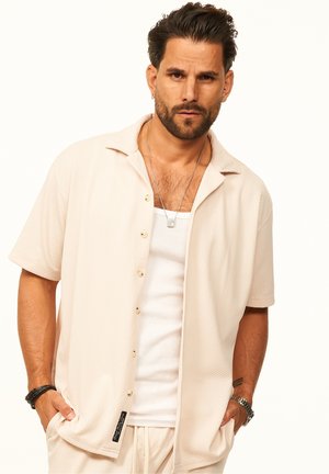 Mann mit gestyltem dunklem Haar und Bart trägt beige kurzärmliges Hemd, weißes Tanktop, Halskette, Uhr und Armbänder, steht mit den Händen in den Taschen.