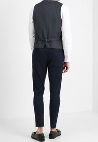 Dopasowany trzyczęściowy garnitur z teksturowaną granatową kamizelką, dopasowanymi granatowymi spodniami i białą koszulą. Całość dopełniają czarne błyszczące mokasyny.