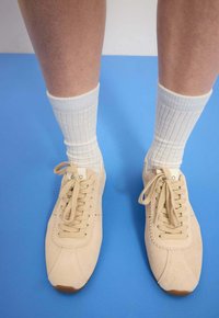 Beżowe zamszowe trampki z zaokrąglonym czubkiem i falistym brzegiem. W połączeniu z jasnymi skarpetkami z prążkowanej bawełny. Na niebieskim tle.