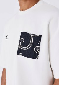 Camiseta blanca con un bolsillo de tela negra estampada y un pequeño logotipo negro en la parte superior izquierda del pecho.
