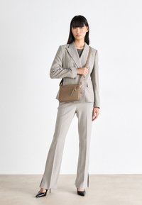 Bruine leren crossbody tas met een ritsvak, gladde textuur en verstelbare riem, gecombineerd met een op maat gemaakt grijs pak en zwarte hakken.