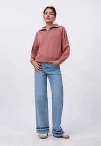 Sweatshirt rosa claro com gola de zip, de mangas longas, combinado com jeans azuis de pernas largas com bainhas desfiadas e sapatilhas.