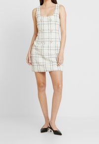 Vit tweed mini-klänning med fyrkantig halsringning, svart rutmönster och sex dekorativa knappar framtill. Matchad med svarta klackar.