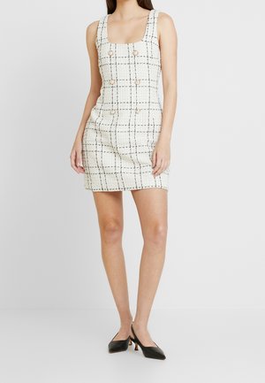 Witte tweed mini-jurk met een vierkante halslijn, zwart ruitpatroon en zes decoratieve knopen aan de voorkant. Gecombineerd met zwarte hakken.