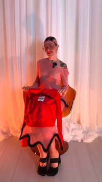 Rød lynlåstrøje med sort kant, vist i hænderne. Modellen har en lyserød ribbet off-shoulder kjole på, sorte platformsko og har synlige tatoveringer.