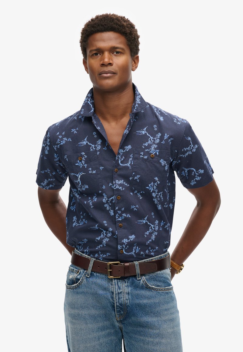 Camisa de botones de manga corta, color azul marino, con un patrón floral en azul claro, bolsillos en el pecho y botones marrones. Llevada con jeans de denim claro y un cinturón.