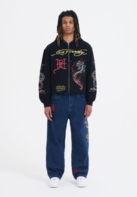 Felpa nera con zip e grafiche multicolori, tra cui una tigre e un serpente. Abbinata a jeans di denim blu larghi con loghi.