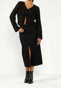 Czarna, dziana, siatkowa krótka kardigan z zapięciem na wstążkę, w zestawie z czarną, wysoką spódnicą dżinsową z rozporkiem z przodu. Błyszczące czarne botki do kostek.