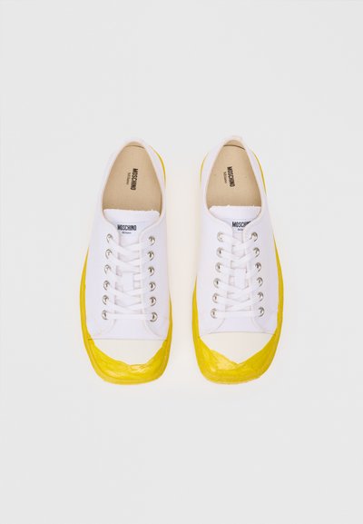 White canvas sneakers with a yellow rubber toe cap. Features include six silver eyelets and a branded insole. Rubber sole for traction.