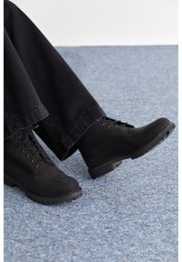 Botas de tobillo de ante negro con suela resistente, con cordones, combinadas con pantalones negros holgados frente a un fondo azul texturizado.