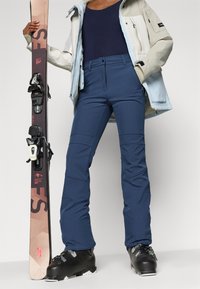 Skibukser i mørkeblått, med en tettsittende design og sømdetaljer. Kombinert med svarte skistøvler og holder ski med bindinger.