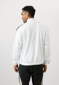 Witte lichtgewicht jas met een opstaande kraag, elastische manchetten en een zwart zijstreep patroon. Soepele textuur, relaxte pasvorm, geschikt voor sportkleding.