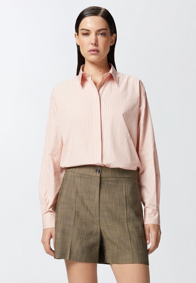 Svijetloružičasta prugasta košulja dugih rukava s kragnom, u kombinaciji s maslinasto zelenim skrojenim šortsima koji imaju prednje dugme i nabore.